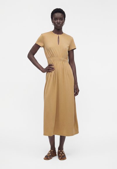 Femme debout, main sur la hanche, portant une robe midi beige à manches courtes et col en forme de trou de serrure, associée à des sandales marron.