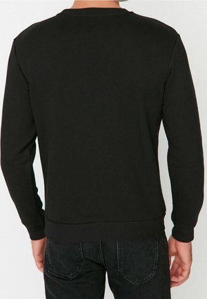 Man met een effen zwart shirt met lange mouwen en zwarte jeans, gezien vanaf de achterkant tegen een neutrale achtergrond.