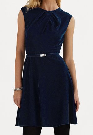 Robe sans manches en velours bleu marine, longueur au genou, avec détail de plissé au niveau du décolleté et fine ceinture à boucle argentée à la taille, portée avec des collants noirs.