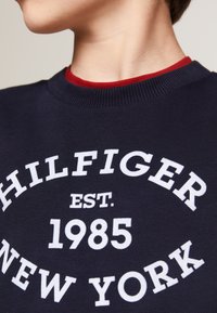 Felpa blu navy con collo a costine rosso; presenta un testo bianco che recita "HILFIGER EST. 1985 NEW YORK." Tessuto liscio con un design casual.