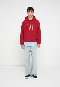 Felpa con cappuccio rossa con logo "GAP" bianco, abbinata a una camicia a righe azzurra chiaro, jeans celesti strappati e sneakers nere.