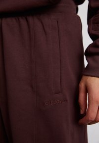 Karmīnu krāsas Adidas sporta bikses ar gludu tekstūru, ar sānu kabatām un diskrētu logo izšuvumu uz augšstilba. Redzams elastīgs jostasvīrs.
