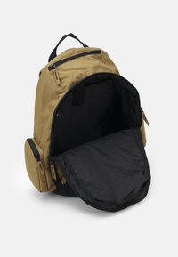 Beżowy plecak z dużym czarnym wnętrzem i zamykanymi kieszeniami po bokach. Posiada regulowane ramiączka oraz uchwyt na górze.
