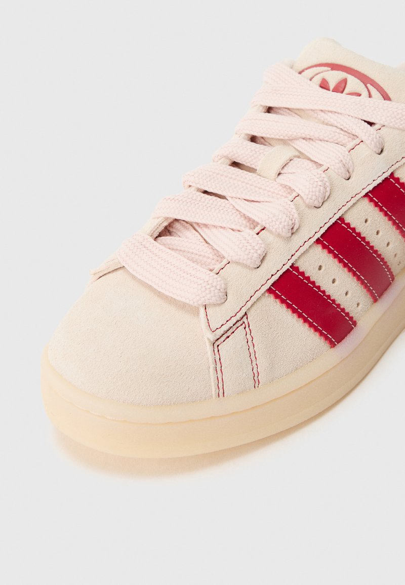 Zapatilla deportiva de ante rosa claro con acentos rojos, tres rayas horizontales y cordones texturizados. Suela plana de goma y detalles sutiles de costura.