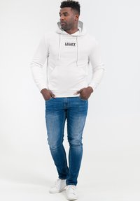 Witte hoodie met "LEGACY" tekst op de borst, kangoeroezak, capuchon met trekkoord, gecombineerd met blauwe spijkerbroek en witte sneakers.