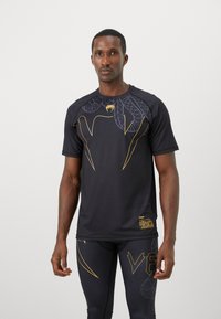 Hombre atlético vestido con una camiseta negra de manga corta y leggings con diseño de serpiente amarilla y el texto parcial "VENUM" en los leggings, de pie contra un fondo blanco.