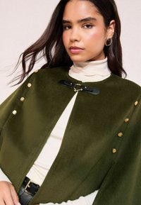 Olivine roheline kapuuts pehme tekstuuriga, kuldsete nuppudega ja musta kunstnahast sulgemisega. Kandu sügise koorevalge turtleneck sviitri peal.