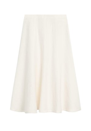 Frau in grauem ärmellosem Oberteil, weißem Midi-Rock und schwarzen Sandalen lehnt an einer weißen Wand und berührt ihr Ohr.