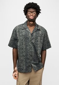 Κοντομάνικο πουκάμισο με σχέδιο δέρματος φιδιού σε αποχρώσεις πράσινου και μαύρου. Διαθέτει γιακά τύπου κάμπινγκ και μία τσέπη στο στήθος.