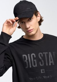 Juodas medvilnės džemperis, ant priekinės pusės turintis didelį iškilų "BIG STAR" tekstą. Derinamas su atitinkančiu juodu kepuraitė, glotnia tekstūra ir minimaliai išpildyta dizaino.