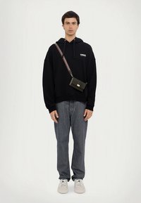 Sweatshirt preta com capuz e gráfico frontal, combinada com jeans azul claro e ténis brancos. Uma mala preta de tiracolo com uma alça às riscas.