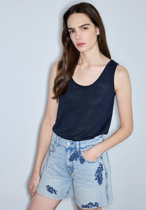 Vrouw met lang bruin haar die een mouwloos zwart topje en lichte spijkershorts met donkerblauwe bloemenborduursel draagt, staande tegen een effen achtergrond.