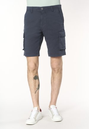 Pantaloncini cargo blu navy con bottone e cerniera, tasche laterali con patta, indossati con scarpe da ginnastica bianche, tatuaggio visibile sul polpaccio sinistro.
