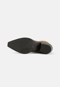 Suela de zapato de ante marrón con una base de goma negra elegante. Presenta una forma de punta afilada y un patrón de agarre texturizado. Logo de marca visible.