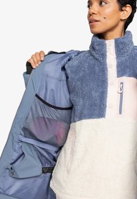 Fleece vest with a blue and cream color scheme, featuring a high collar, front zipper, and mesh pocket inside. Soft texture and layered design.