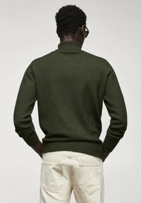 Dunkelgrüner Strickpullover mit einem gerippten Kragen und Bündchen, der ein strukturiertes Muster aufweist, kombiniert mit hellfarbigen Hosen.