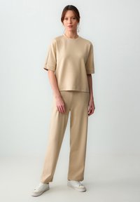 Beżowy dwuczęściowy zestaw do wypoczynku, zawierający bluzkę z krótkim rękawem i okrągłym dekoltem o luźnym kroju oraz szerokie spodnie. Miękka faktura.