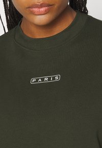 Musta vihreä ribattu collegepaita, jossa on pyöreä kaula-aukko ja edessä valkoinen "PARIS"-logo brodeerattuna. Pehmeä, teksturoitu kangas.