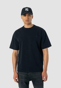 Mies, der eine schwarze New York Yankees Cap, ein schwarzes Übergröße-T-Shirt und dunkle Jeans trägt, steht vor einem schlichten grauen Hintergrund.