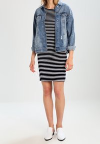 Veste en jean avec une finition bleu délavé, portée par-dessus une robe à rayures noir et blanc. Le modèle porte des chaussures blanches à enfiler. Design simple.