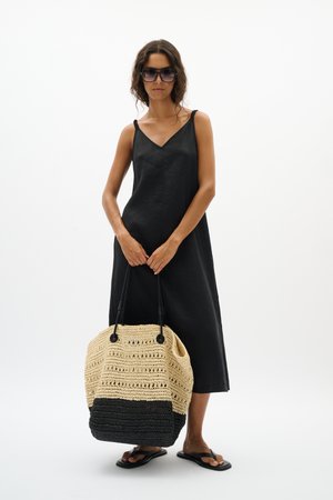 Femme portant une robe noire sans manches et des sandales, tenant un grand sac fourre-tout tissé beige et noir, avec des lunettes de soleil sur fond blanc.