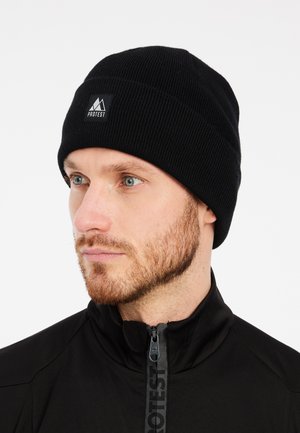 Berretto lavorato a maglia nero con una texture a coste e un piccolo patch con il logo "PROTEST" in nero sulla parte anteriore, indossato da un modello con la barba.