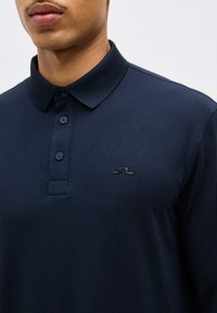 Άνδρας που φοράει μακρυμάνικη μπλε ναυτική πόλο μπλούζα με τρία κουμπιά και ένα μικρό μαύρο λογότυπο στο αριστερό στήθος.