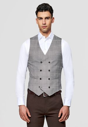 Mężczyzna ubrany w szarą, dwurzędową kamizelkę w kratę na białej koszuli i brązowe spodnie, stojący na jednolitym tle.