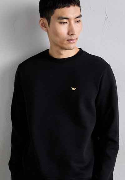 Black sweatshirt with a round neckline, long sleeves, and a small gold logo on the chest. Smooth texture and a relaxed fit.