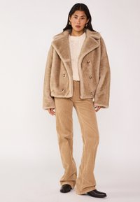 Manteau en fausse fourrure à double boutonnage de couleur marron clair avec large col, associé à un pantalon flare en velours côtelé beige et des bottines noires.