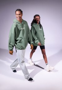 To modeller iført matchende store grønne hettegensere med frontlomme og hvit logo. Den ene modellen har på seg lyse denimbukser, mens den andre har på seg svarte shorts.
