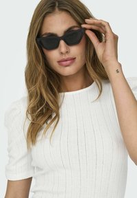Sorte cat-eye solbriller med mørke linser; mat overflade. Båret af en model med langt, bølget hår, iført en struktureret hvid top med korte ærmer.