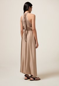 Beige satiini maksimekko, jossa on halterneck-olkaimet, ker gathered vyötärö ja pitkät takanauhat. Sileä rakenne. Yhdistetty mustiin hihnallisiin sandaaleihin.