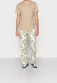 Beige short-sleeve t-shirt paired with white floral patterned trousers with green accents. Model wears green and white sneakers.