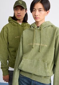 Du modeliai alyvuogių žalios spalvos džemperiuose su vintage apdaila ir priekinėmis kišenėmis, dekoruoti auksine siuvinėta "ALPHA INDUSTRIES" užrašu.