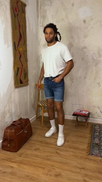 Νέος άνδρας με dreadlocks στέκεται σε φθαρμένο δωμάτιο φορώντας λευκό μπλουζάκι, τζιν σορτς, λευκά αθλητικά κοντά σε καρέκλα, δερμάτινη τσάντα, πολύχρωμο τοίχο και βιβλία.