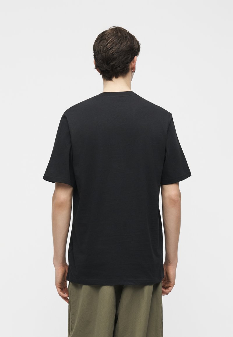 Person mit kurzen braunen Haaren, die ein schlichtes schwarzes T-Shirt mit kurzen Ärmeln und olivgrüne Hosen trägt, von hinten vor weißem Hintergrund gesehen.