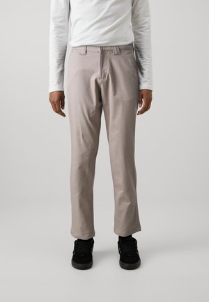 Lichtgrijze broek met een gladde textuur, rechte pasvorm en twee voorkwakken. Ggedragen met een wit overhemd met lange mouwen en zwarte sneakers.
