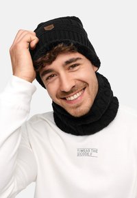 Black knitted beanie with a folded cuff and a brown logo patch, paired with a matching ribbed neck warmer, worn over a white sweatshirt.