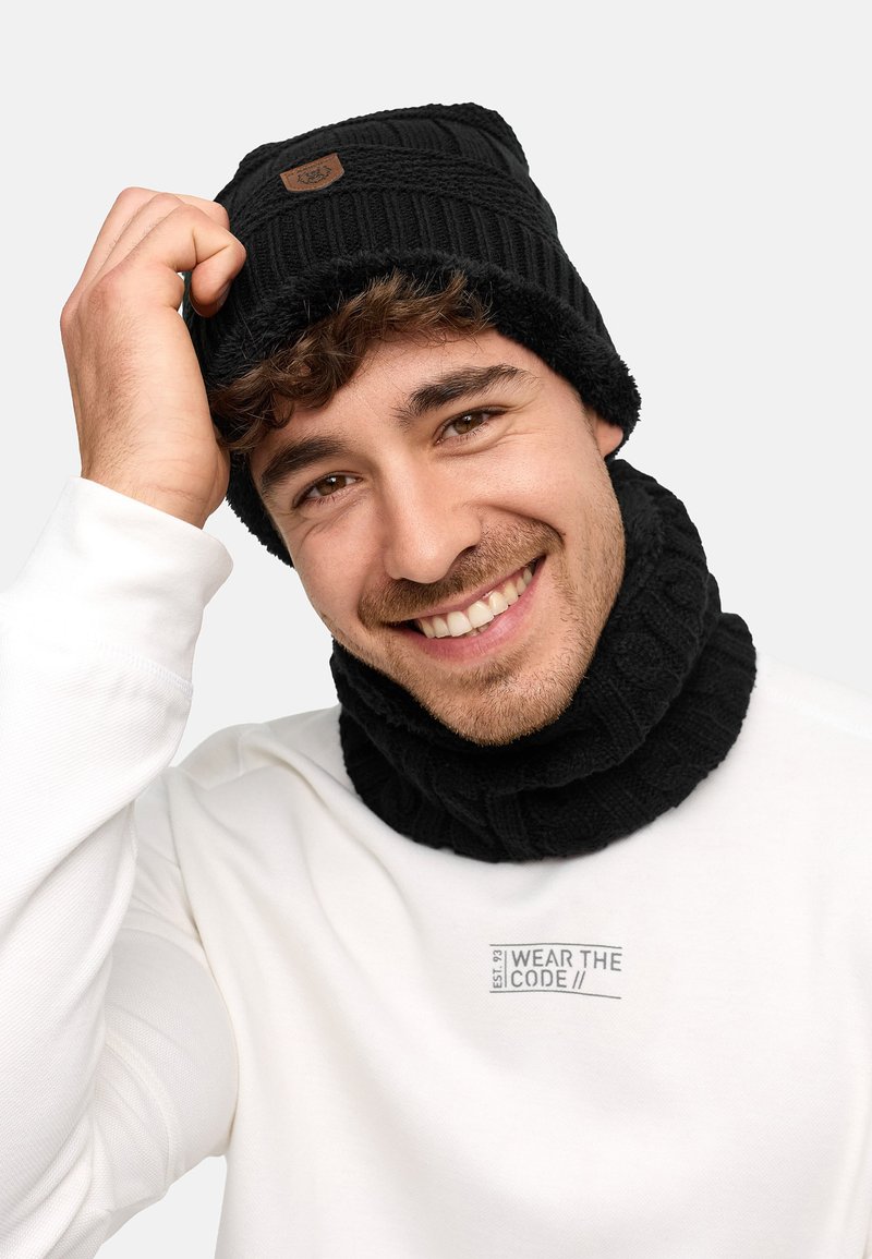 Black knitted beanie with a folded cuff and a brown logo patch, paired with a matching ribbed neck warmer, worn over a white sweatshirt.