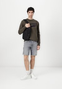 Crni crossbody torba, maslinasto zeleni dukserica s logom, svijetloplave traper kratke hlače, bijele tenisice, stoji pred jednostavnom pozadinom.