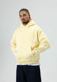 Geel katoenen hoodie met een buidelzak, geribbelde boorden en zoom. Heeft een klein logo op de voorkant en wordt gedragen met donkere jeans.