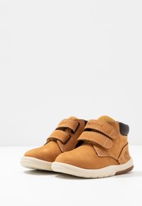 Brown suede shoes with double Velcro straps, rounded toes, and contrasting white and brown soles; features textured stitching and logo.