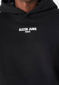 Sudadera negra de tela suave con un estampado frontal en blanco que dice "SIXTH JUNE PARIS." El diseño incluye una capucha con cordón.