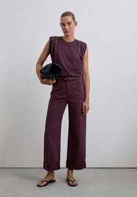Un haut à manches courtes et un pantalon large en bourgogne profond. Le haut est orné d'accents à clous aux épaules. Une pochette noire et des sandales plates complètent le look.