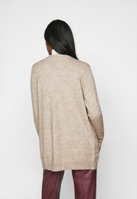 Beige kardigán hosszú ujjal, laza fazonnal, bordázott mandzsettákkal és szegéllyel. Puha tapintás, nincs látható minta, bordó nadrág fölött viselve.