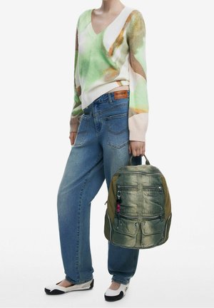 Suéter con patrón verde y beige, jeans azul sueltos y una mochila verde texturizada con varios bolsillos con cremallera, sostenida en una mano.