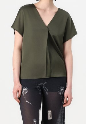 Frau trägt eine olivgrüne Bluse mit V-Ausschnitt und kurzen Ärmeln sowie schwarze transparente Hosen mit weißen Blumen- und Feendesigns, steht vor einem einfarbigen Hintergrund.