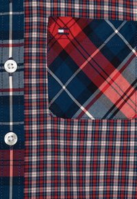 Zbliżenie na koszulę w kratę w kolorach czerwonym, białym i niebieskim, z kieszonką na piersi oraz białymi guzikami z logo "Tommy Hilfiger."
