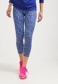 Niebieskie legginsy w wzory z teksturowanym designem, o długości do połowy łydki, połączone z jasnoróżowymi butami sportowymi.